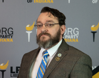 Portrait of Evan McMahon, wearing a brown tweed jacket, white collar shirt, and a blue and yellow striped tie.